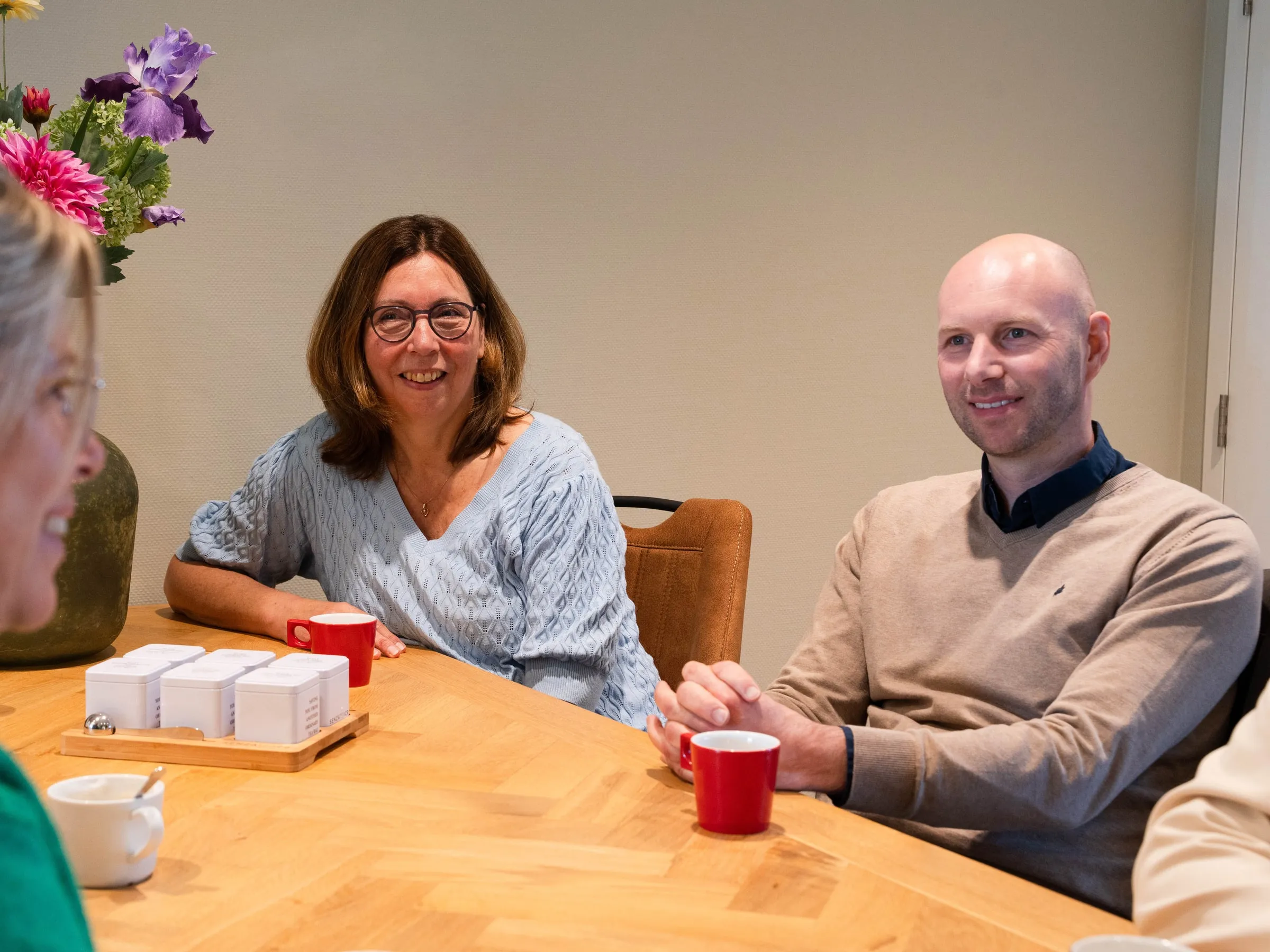 Drie collega's in overleg aan een houten tafel met koffie; twee van hen luisteren naar de persoon die op de voorgrond zit.